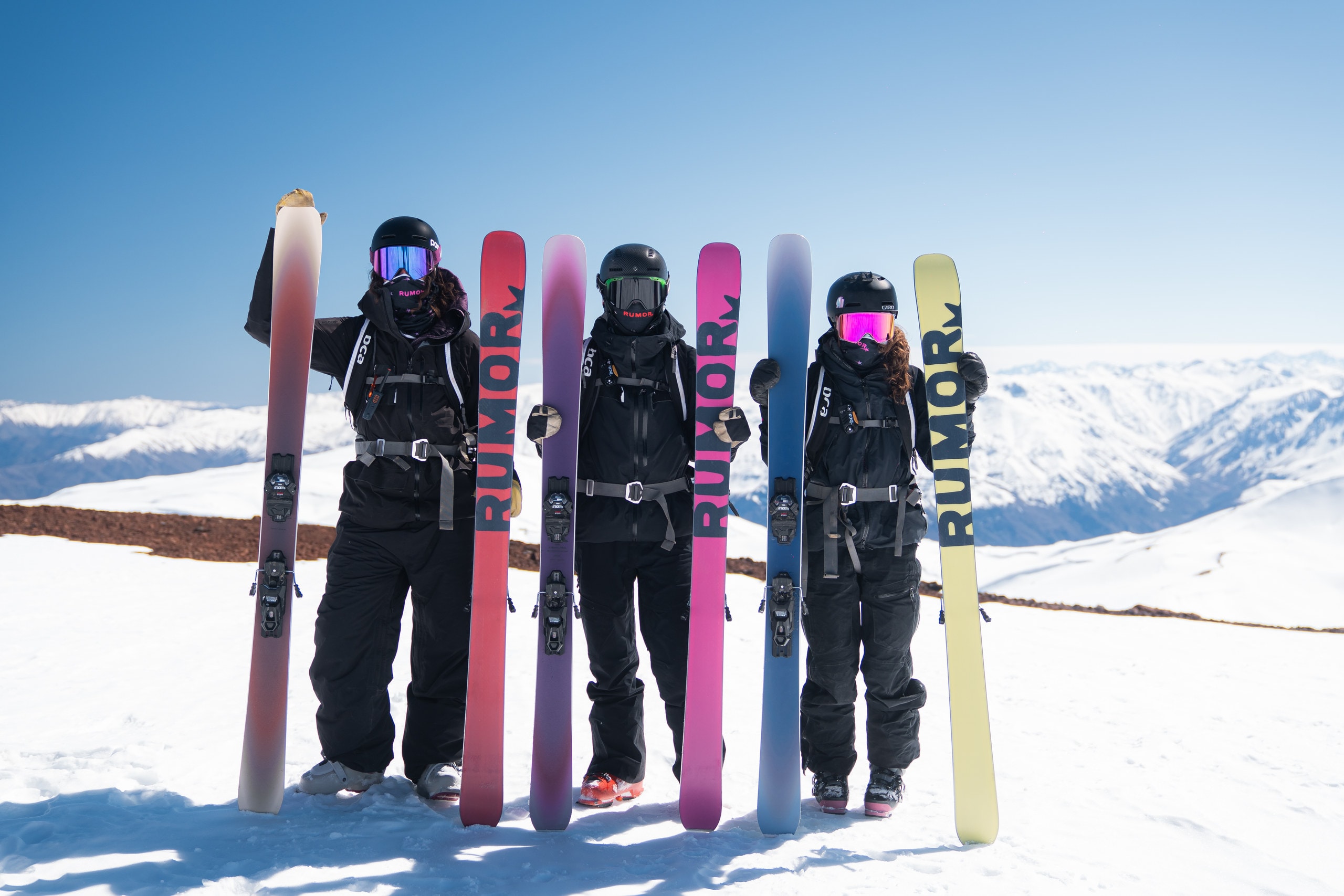Three skiers standing on a snowy ridge, each holding up a pair of RUMOR skis in a different colorway