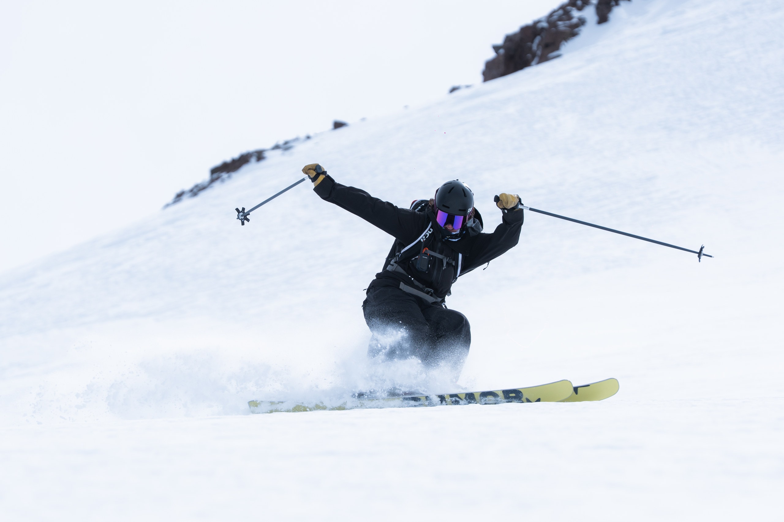A skier leaning into a carve, snow spraying off a pair of yellow RUMOR skis